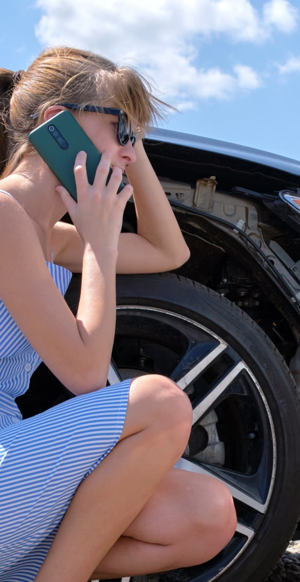 A woman crouched by her car outside while talking on her phone